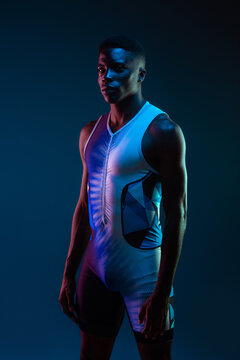 Side view of serious concentrated young African American male athlete standing against blurred blue neon background in dark studio looking at camera