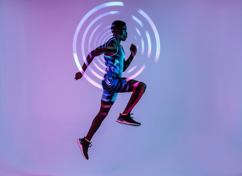 Side view of energetic young African American male sprinter jumping against purple wall with concentric circles in studio with neon lights neon background
