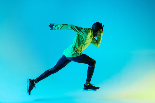 Full body side view of young African American male runner in colorful tracksuit sprinting from start position in studio with bright neon illumination