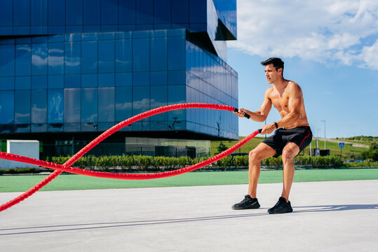 Side view of muscular male athlete with naked torso doing exercises with battle ropes on terrace during sunny day in summer
