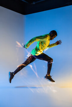 Full body side view of young African American male runner in colorful tracksuit sprinting from start position in studio with bright neon illumination