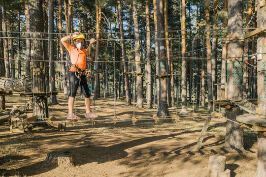 Brave child in safety helmet and harness walking along suspension bridge in adventure park while enjoying weekend and looking at camera