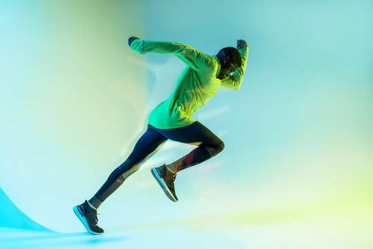 Full body side view of young African American male runner in colorful tracksuit sprinting from start position in studio with bright neon illumination