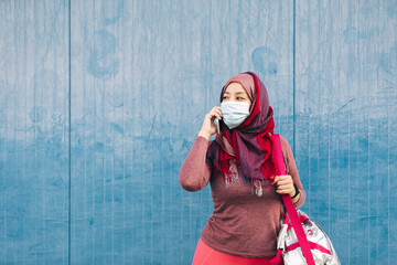Arab female in sportswear and medical mask standing near building in city speaking on mobile phone after training while looking away