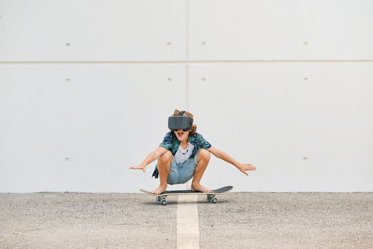 Boy in summer clothes skating on skateboard wearing virtual reality glasses and gesturing with the arms raised on the street
