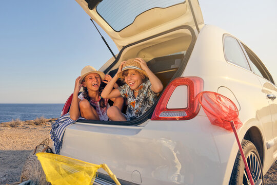 Two children sitting inside the trunk of the car making jokes while touching the straw hat with baskets of objects for the beach next to the car with the sea in the background