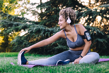 Happy young ethnic female with curly hair in sportswear listening to music with headphones and smartphone and doing seated forward bend exercise while stretching body during fitness training in summer park