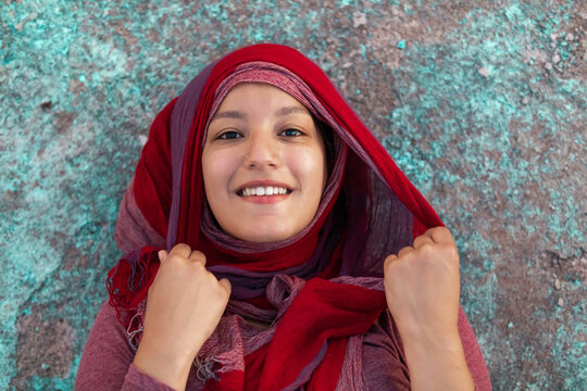 Happy Arab female wearing hijab leaning on concrete colorful building in city smiling at camera
