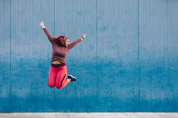 Carefree ethnic female wearing hijab celebrating victory and jumping with raised arms near building with blue wall while looking away