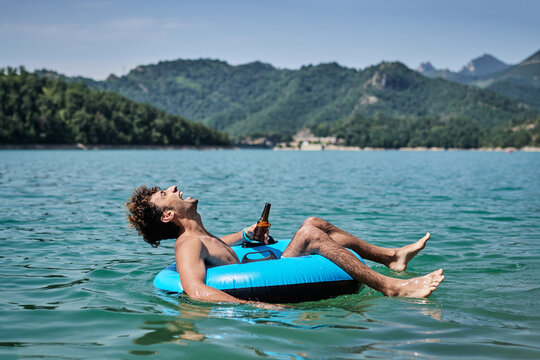 Content male with bottle of beer floating in inflatable ring on surface of calm water of pond while enjoying sunny day in summer and smiling