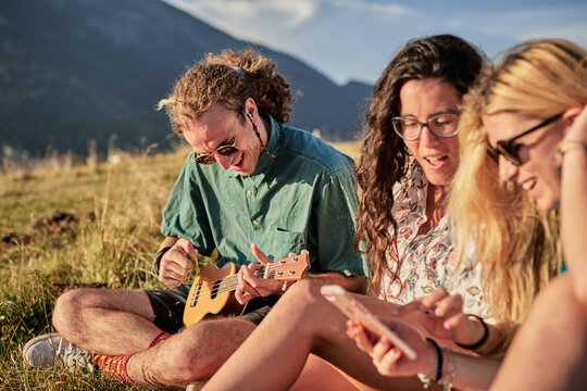 Cheerful friendly people playing ukulele and browsing smartphones while entertaining during weekend in summer and sitting on grass in highlands