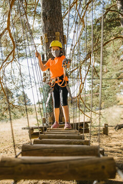 Concentrated Kid In Medical Mask And Safety Equipment Standing On Wooden Suspension Bridge In Adventure Park During COVID 19 Epidemic