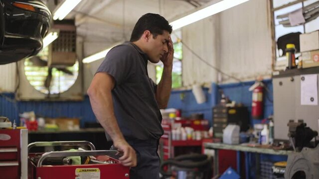 Hispanic car mechanic working in auto shop, having migraine headache, using mobile phone to check in with tele-medicine doctor  - Powered by Adobe