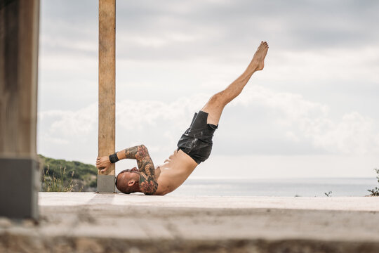 Side view of muscular male in shorts performing shoulder stand while leaning on wooden column during training