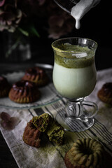 Pouring foamed milk into matcha latte in glass with matcha tea powder. Matcha latte with mini chocolate-matcha bundt cakes and hydrangea flowers on dark background.