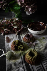 Mini chocolate-matcha bundt cakes, bowl with chocolate and hydrangea flowers on linen towel on dark background.