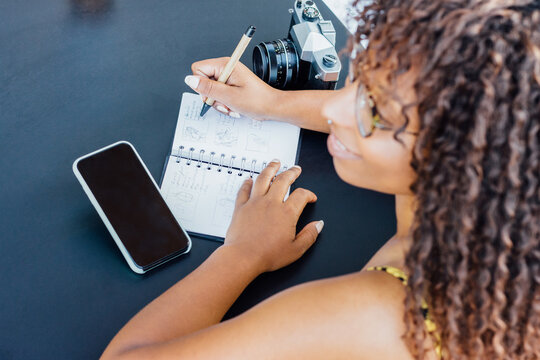 From above woman sitting at table in cafe and working remotely while writing in notepad