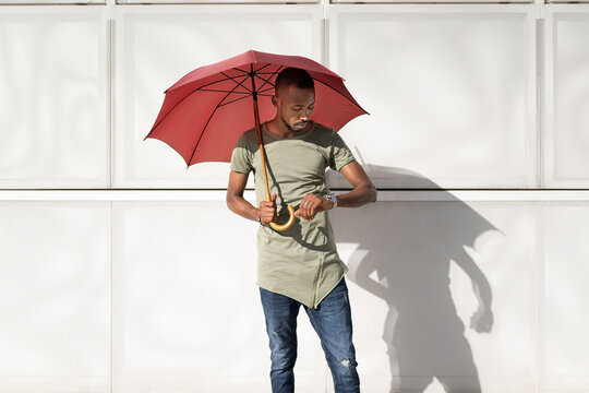 Serious African American male standing under umbrella on street on sunny day and checking time on wristwatch