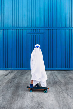 Child in ghost costume standing on skateboard in city during Halloween