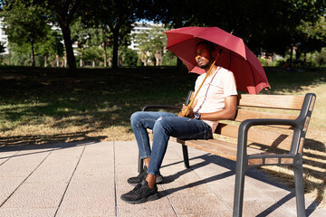 Peaceful African American male sitting under umbrella on wooden bench in park and enjoying weather on sunny day in summer