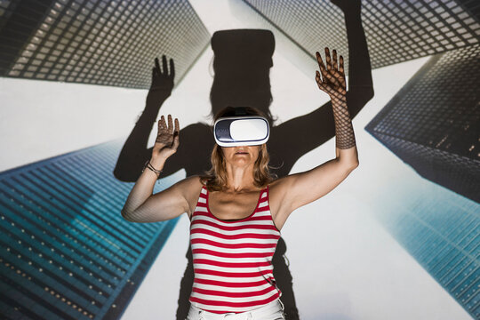Unrecognizable female in casual wear and VR headset standing with arms raised against contemporary skyscrapers inside virtual city