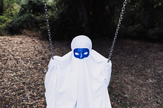Unrecognizable kid in white ghost costume swinging and playing on playground during Halloween holiday