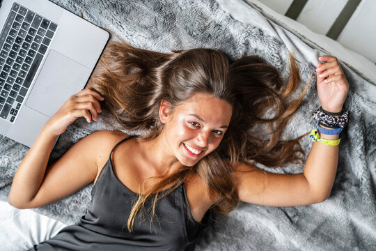 Top view of delighted female in sleepwear lying on cozy bed at home and typing on netbook in morning while chatting on social media looking at camera