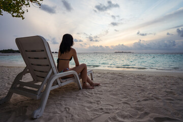 Back view of unrecognizable female tourist in bikini sitting on chair and enjoying  amazing sunset over waving sea while resting on sandy beach during summer vacation on Maldives islands