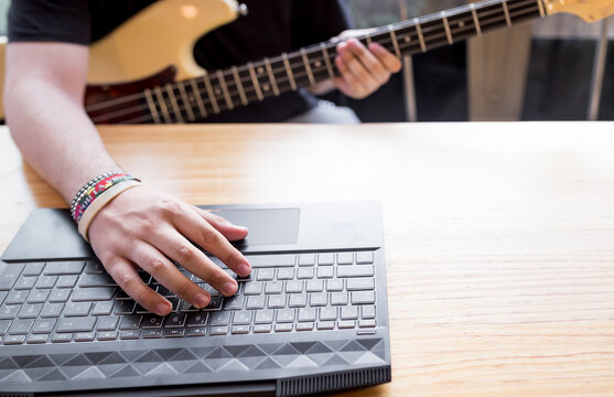 Crop young male sitting at table and watching video tutorial on laptop while learning to play music on bass guitar