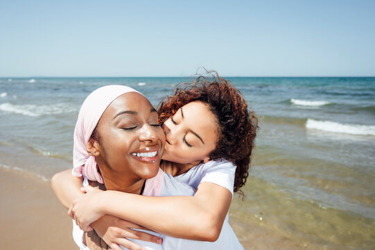 Delighted black mother piggybacking young daughter while having fun with closed eyes at seaside in summer