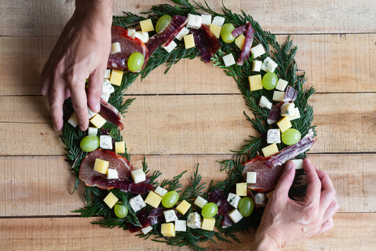 Top view of hands placing creative Christmas wreath made with green herbs and decorated with fresh grape and chopped cheese and ham arranged on wooden table