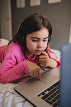 Focused little girl in headphones lying on bed and watching online video on laptop while spending free time at home