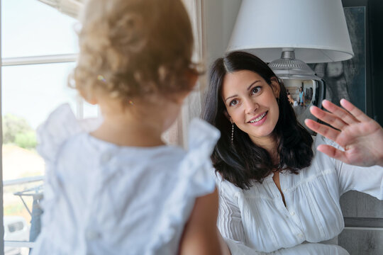 Mother waving to a little girl in a white dress sitting on the floor of a dining room in a house
