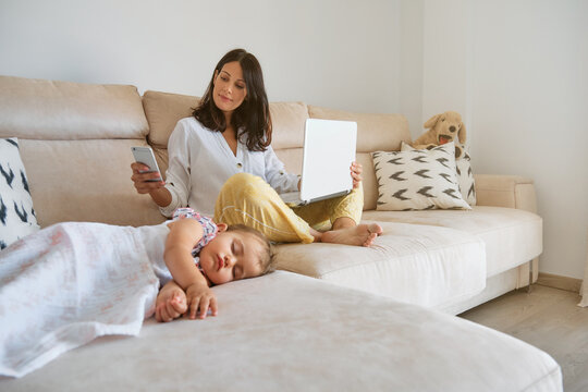 Little girl sleeping on the sofa while her mother works with a laptop and using the mobile next to her