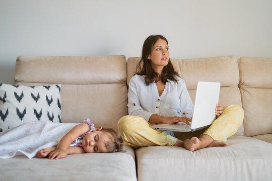 Woman working on the sofa with distracted expression with the laptop while a toddler is sleeping on a side at home