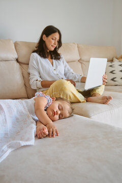 Little girl sleeping on the sofa while her mother works with a laptop next to her