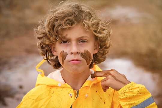 Blond kid with curly hair in a yellow raincoat drawing marks on his face with the mud looking at the camera with serious expression in the middle of the forest