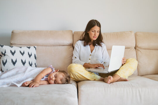 Woman working on the sofa with the laptop while a toddler is sleeping on a side at home