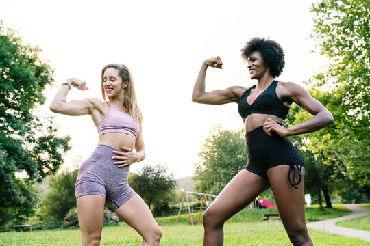 Low angle of happy young fit women in sportswear flexing muscles while exercising together on green lawn in park