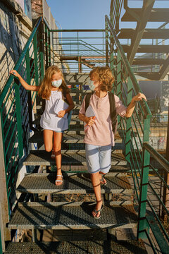 Vertical photo of two children wearing masks and carrying school bags down an outside staircase with homework in hand