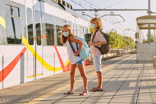 Two children with black school bags and mask on the platform of a station with the train stopped on the track in the morning