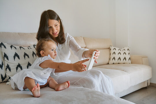 Little girl in a white dress pointing to a smartphone held by her mother sitting on the sofa of a house
