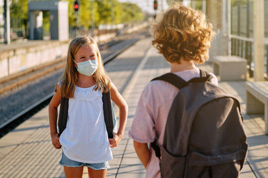 Two children with black school bags and mask talking from a distance while waiting for the train on a station platform in the morning