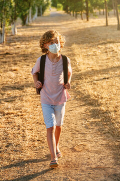 Vertical photo of the portrait of a blond boy with curly hair wearing a mask and school bag while walking along a path surrounded by trees