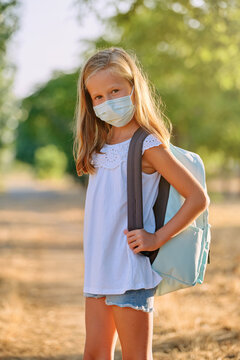 Vertical photo of a portrait of a blonde girl with a mask and a school bag looking at a camera in the middle of nature