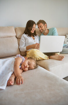 Little girl sleeping on the couch while a pregnant couple is distracted with a laptop next to her at home
