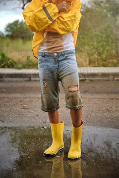 Vertical photo of the detail of the body of a boy in a yellow raincoat and yellow rain boots standing in the middle of a puddle in a road on a rainy day