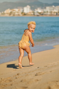 Vertical photo of a girl in a yellow and white striped swimsuit with a hair band running down to the seashore and turning to look to her back on a sunny day