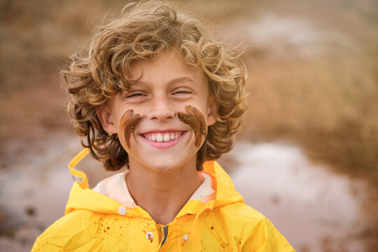 Blond kid with curly hair in a yellow raincoat with marks on his face with the mud looking at the camera with serious expression in the middle of the forest