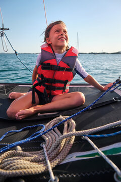 Vertical photo of a blond boy with life jacket sitting on the canvas of a boat in the middle of the sea looking at the sky with the seashore in the background
